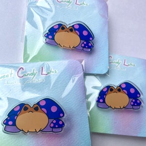 May include: Several pins featuring a cartoon frog with a mushroom cap. The frog is tan with a grumpy expression. The mushroom caps are blue with pink polka dots. The pins are packaged on a pastel gradient card with the text "Sweet Candy Lotus".