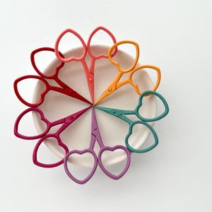 May include: A set of eight heart-shaped scissors in various colours, including pink, red, orange, yellow, green, and purple, arranged in a circle on a white plate.