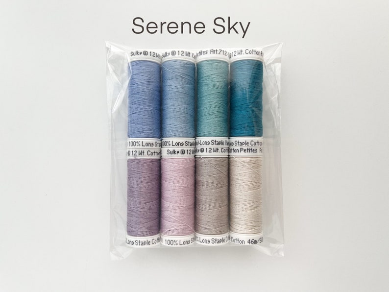 May include: A set of six spools of thread in shades of blue, purple, pink, gray, and white. The thread is labeled "Sulky 12 Wt. Cotton" and "Long Staple Cotton".