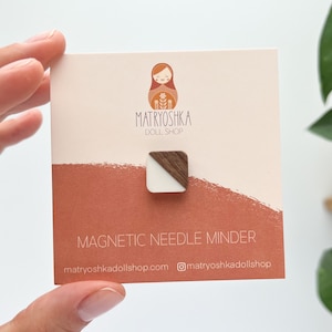 以下が含まれることがあります： 「MATRYOSHKA DOLL SHOP」の文字とウェブサイトアドレス「matryoshkadollshop.com」、Instagramのハンドル「@matryoshkadollshop」が書かれた白と茶色の木製磁気針止め。