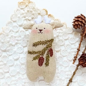 May include: A handmade beige linen reindeer ornament with embroidered pine branches and brown pine cones. The ornament is sitting on a white lace doily.