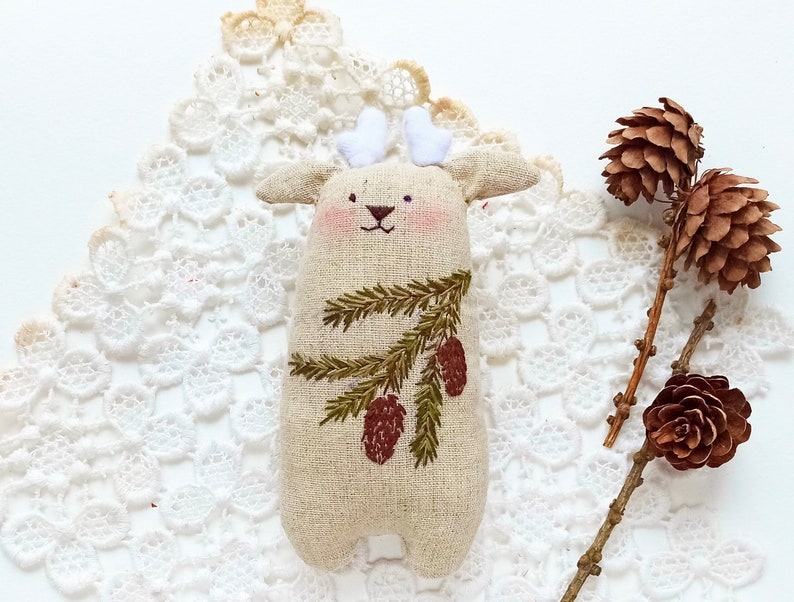 May include: A handmade beige linen reindeer ornament with embroidered pine branches and brown pine cones. The ornament is sitting on a white lace doily.