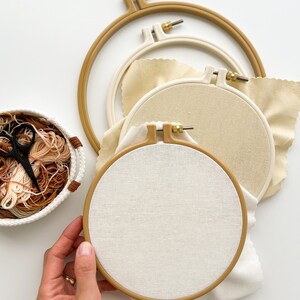 May include: Three embroidery hoops with beige fabric, one of which is being held by a hand. The hoops are nested inside each other, with the largest hoop on the bottom. A basket of embroidery floss is in the background.