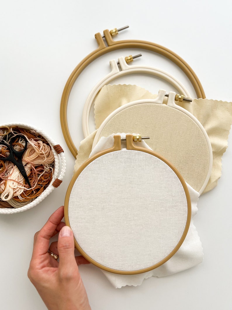 May include: Three embroidery hoops with beige fabric, one of which is being held by a hand. The hoops are nested inside each other, with the largest hoop on the bottom. A basket of embroidery floss is in the background.
