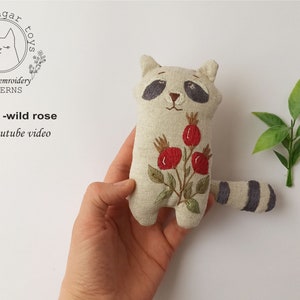 May include: A hand holds a beige linen raccoon plushie with a gray striped tail. The raccoon has embroidered black eyes, a black nose, and a red wild rose with green leaves on its chest. The text "raccoon -wild rose" and "PDF+youtube video" is visible below the plushie.