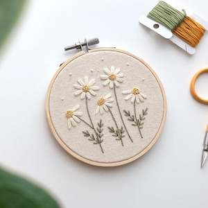 Puede incluir: Un aro de bordado blanco con un dise&ntilde;o floral de margaritas blancas con centros amarillos y tallos verdes. El aro est&aacute; sobre una superficie blanca con un par de tijeras de bordado y dos carretes de hilo de bordar.