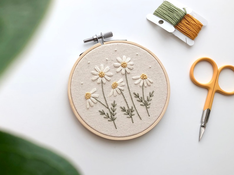 Puede incluir: Un aro de bordado blanco con un dise&ntilde;o floral de margaritas blancas con centros amarillos y tallos verdes. El aro est&aacute; sobre una superficie blanca con un par de tijeras de bordado y dos carretes de hilo de bordar.