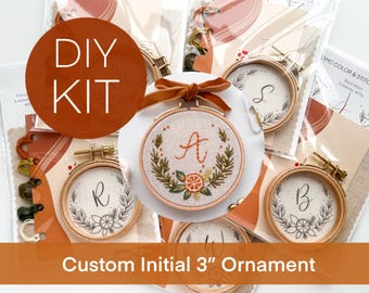 May include: DIY kit for a custom initial 3-inch ornament. The image shows several embroidery hoops with letters, surrounded by floral designs. The text "DIY KIT" is in a circle. The ornament has a brown velvet ribbon.