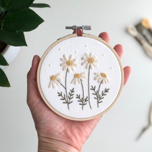 Puede incluir: Un aro de bordado blanco con un dise&ntilde;o floral de margaritas blancas con centros amarillos y tallos verdes. El aro se sostiene en una mano.