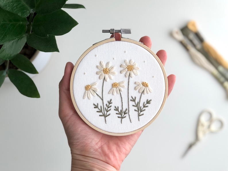 Puede incluir: Un aro de bordado blanco con un dise&ntilde;o floral de margaritas blancas con centros amarillos y tallos verdes. El aro se sostiene en una mano.
