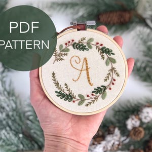 May include: Embroidered hoop art featuring a floral wreath with the letter "A" in gold thread. The design includes green leaves and red berries. The text "PDF PATTERN" is in a green circle.