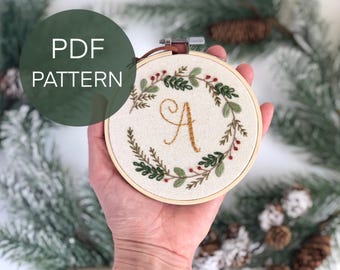 Peut inclure: Broderie sur tambour avec une couronne florale et la lettre "A" en fil doré. Le motif comprend des feuilles vertes et des baies rouges. Le texte "PDF PATTERN" est dans un cercle vert.