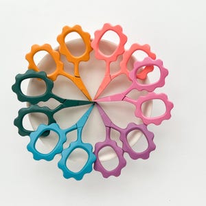 May include: A set of eight colorful flower-shaped scissors arranged in a circle on a white surface. The scissors are in shades of orange, pink, teal, and purple.