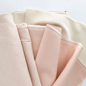 May include: A stack of four fabric swatches in various shades of pink and cream. The fabric is smooth and has a subtle sheen.