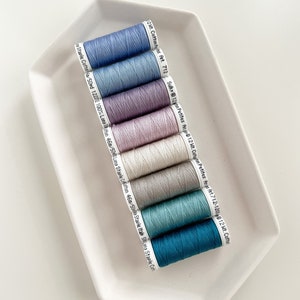 May include: Eight spools of thread in various shades of blue, purple, and white. The spools are labeled with the brand name "Sulky" and the weight of the thread.