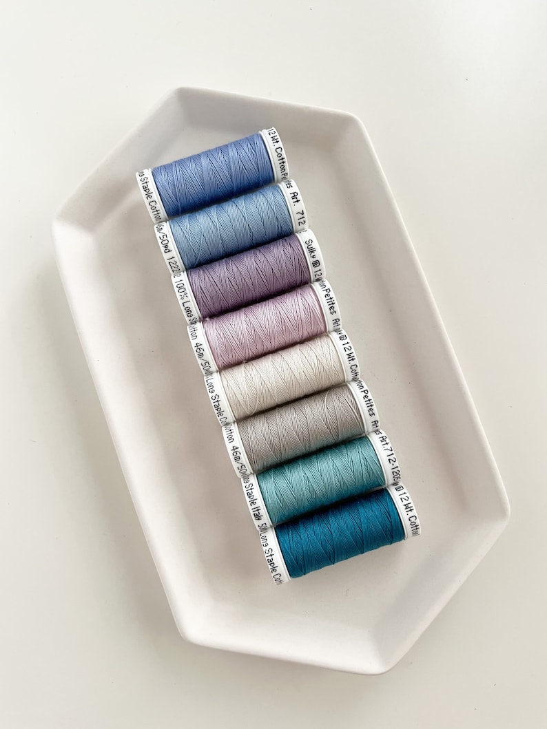 May include: Eight spools of thread in various shades of blue, purple, and white. The spools are labeled with the brand name "Sulky" and the weight of the thread.