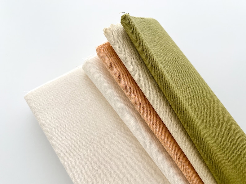 May include: A stack of five fabric swatches in different colors and textures. The top swatch is a dark green, the next is a light green, followed by a beige, then a light orange, and finally a white swatch.