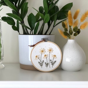 Puede incluir: Un aro de bordado blanco con un dise&ntilde;o floral de margaritas blancas con centros amarillos y tallos verdes. El aro est&aacute; colgado de una correa de cuero marr&oacute;n. El aro est&aacute; sentado en un estante blanco con una planta en maceta y un jarr&oacute;n de flores secas.