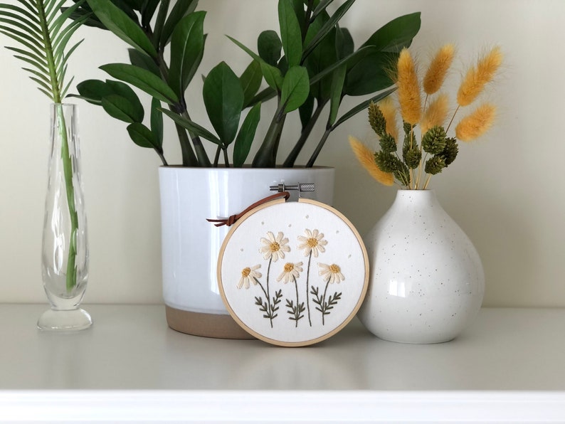 Puede incluir: Un aro de bordado blanco con un dise&ntilde;o floral de margaritas blancas con centros amarillos y tallos verdes. El aro est&aacute; colgado de una correa de cuero marr&oacute;n. El aro est&aacute; sentado en un estante blanco con una planta en maceta y un jarr&oacute;n de flores secas.