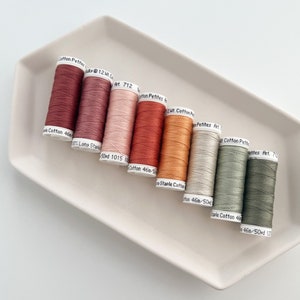 May include: Eight spools of thread in various shades of red, orange, beige, and green. The spools are labelled with the text "Cotton Petites Art. 712", "Cotton 46a-50d 1015", "Cotton 46a-50d", "Staple Cotton 46a-50d", "Cotton Petites Art. 712", "Cotton Petites Art. 712", "Cotton Petites Art. 712", and "Cotton 46a-50d 127".