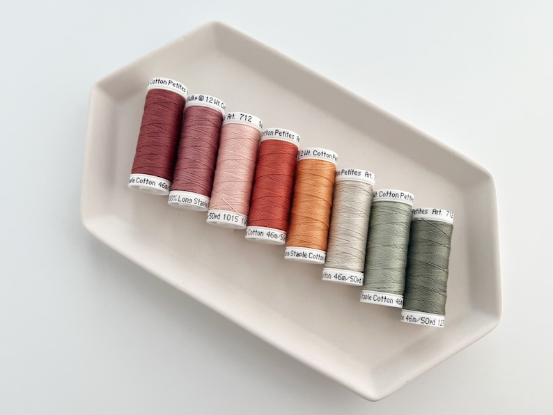 May include: A collection of eight spools of thread in various shades of red, orange, beige, and green. The spools are labeled with the text "Cotton Petites Art. 712", "Cotton 46a-50d 1015", "Cotton 46a-50d", "Staple Cotton 46a-50d", "Cotton Petites Art. 712", "Cotton Petites Art. 712", "Cotton Petites Art. 712", and "Cotton 46a-50d 127".
