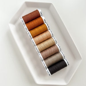 May include: Eight spools of thread in various shades of brown, beige, and black. The spools are labeled with the brand name "Sulky" and the weight of the thread.