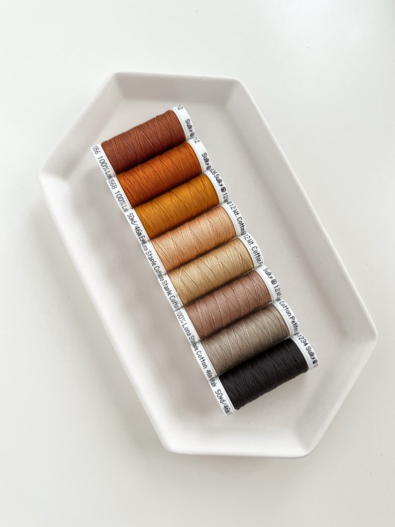 May include: Eight spools of thread in various shades of brown, beige, and black. The spools are labeled with the brand name "Sulky" and the weight of the thread.