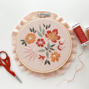 May include: A close-up of a completed embroidery hoop with a floral design featuring orange, pink, and red flowers and leaves. Two spools of embroidery floss are visible in the background, one orange and one red.