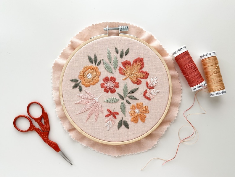 May include: A close-up of a completed embroidery hoop with a floral design featuring orange, pink, and red flowers and leaves. Two spools of embroidery floss are visible in the background, one orange and one red.
