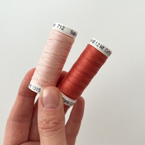 May include: Two spools of thread, one is a light pink color and the other is a burnt orange color. The spools are labeled with the text "12 Wt. Cotton" and the numbers "712" and "1015".