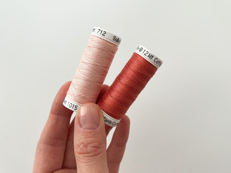 May include: Two spools of thread, one is a light pink color and the other is a burnt orange color. The spools are labeled with the text "12 Wt. Cotton" and the numbers "712" and "1015".