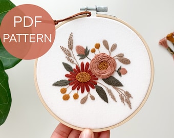May include: Embroidered floral hoop art featuring a pink rose, red daisy, and green leaves. The hoop is made of wood and has a white fabric background.