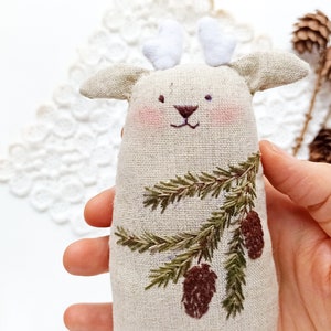 May include: A beige linen deer ornament with embroidered pine branches and pine cones. The deer has white felt antlers.