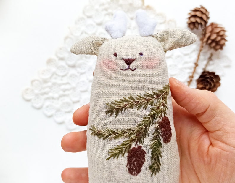 May include: A beige linen deer ornament with embroidered pine branches and pine cones. The deer has white felt antlers.
