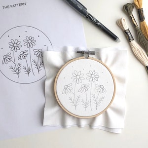 Puede incluir: Un aro de bordado blanco con un dibujo simple de cinco margaritas en su interior. El aro est&aacute; sobre un fondo de tela blanca. El patr&oacute;n para el bordado est&aacute; en una hoja de papel blanca con las palabras "THE PATTERN" en la parte superior.