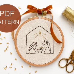 Peut inclure: Un cercle à broder avec un tissu beige représentant un dessin au trait noir d'une scène de la Nativité. Le cercle est orné d'un cadre en bois et d'un ruban de velours marron. Le texte "PDF PATTERN" est dans un cercle marron.