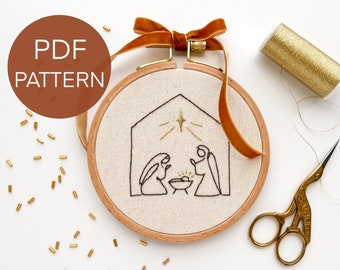 May include: An embroidery hoop with a beige fabric featuring a black line drawing of a nativity scene. The hoop is trimmed with a wooden frame and a brown velvet ribbon. The text "PDF PATTERN" is in a brown circle.