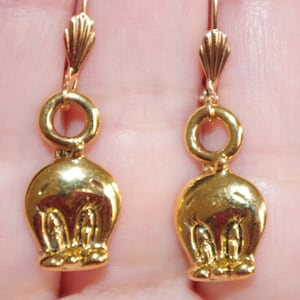 Op de afbeelding: Een paar goudkleurige oorbellen met een cartoonkarakterontwerp. De oorbellen hebben een haaksluiting en een kleine, ronde lus die de haak verbindt met de karakterhanger. De hanger heeft een gestileerde, afgeronde vorm met gezichtskenmerken.
