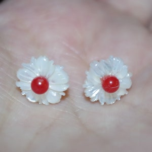 May include: A pair of flower-shaped earrings. Each earring features a white, petal-like design surrounding a vibrant red center. The earrings are small and delicate, with a polished finish, suggesting they are made of a lustrous material.