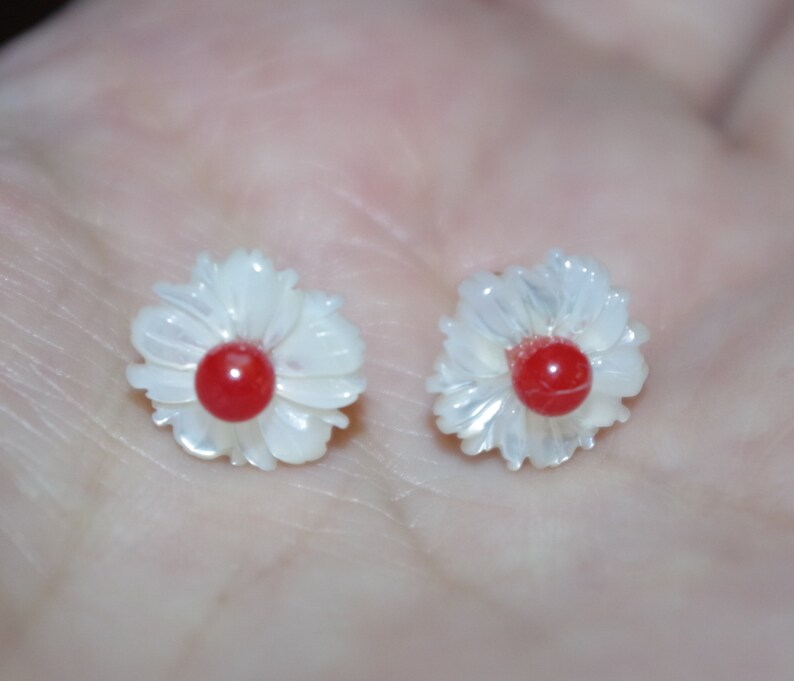 May include: A pair of flower-shaped earrings. Each earring features a white, petal-like design surrounding a vibrant red center. The earrings are small and delicate, with a polished finish, suggesting they are made of a lustrous material.