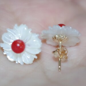 May include: A pair of flower-shaped stud earrings. Each earring features a white, petal-like design surrounding a red center. The posts and backing are gold-colored. The earrings are designed to resemble daisies.