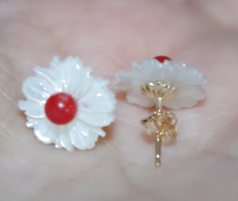 May include: A pair of flower-shaped stud earrings. Each earring features a white, petal-like design surrounding a red center. The posts and backing are gold-colored. The earrings are designed to resemble daisies.