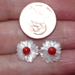 May include: A pair of daisy-shaped stud earrings. The earrings have white, iridescent petals and a bright red center. A penny is next to the earrings for size comparison.