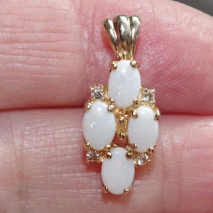 May include: A gold pendant featuring four oval-shaped white opals arranged in a vertical cluster, accented by small clear round gemstones set between the opals. The pendant includes a fluted gold bail at the top for attachment to a necklace chain.