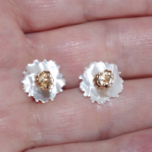 May include: A pair of flower-shaped stud earrings. Each earring features a white, petal-like design surrounding a gold-colored center. The earrings are small and delicate, suitable for everyday wear.