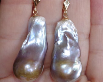 May include: A pair of drop earrings featuring large, irregular baroque pearls with a lustrous, iridescent surface displaying shades of lavender, silver, and gold. The pearls are suspended from polished gold-tone metal lever-back hooks.
