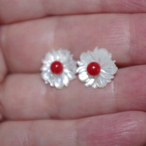 May include: A pair of flower-shaped stud earrings. Each earring features a white, petal-like design surrounding a small, round, red center. The earrings are small and delicate, suitable for everyday wear.