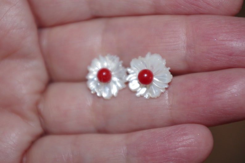 May include: A pair of flower-shaped stud earrings. Each earring features a white, petal-like design surrounding a small, round, red center. The earrings are small and delicate, suitable for everyday wear.