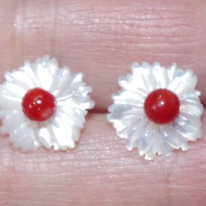 May include: A pair of flower-shaped earrings. Each earring features a white, petal-like design surrounding a vibrant red center. The earrings are likely made of mother-of-pearl and coral, creating a delicate and eye-catching accessory.