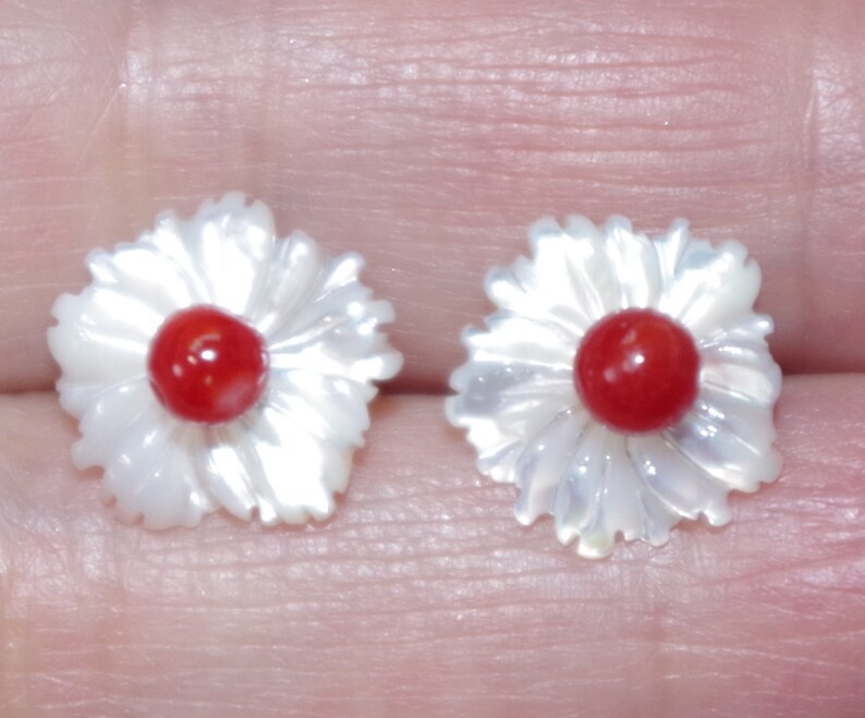 May include: A pair of flower-shaped earrings. Each earring features a white, petal-like design surrounding a central, round, red bead. The earrings are set against a neutral background.